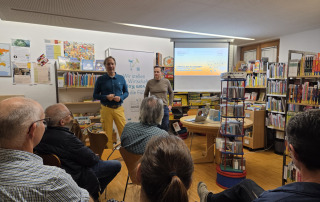 zwei stehende Menschen die sprechen - fünf sitzende Menschen von hinten die zuhören. Vortragssetting
